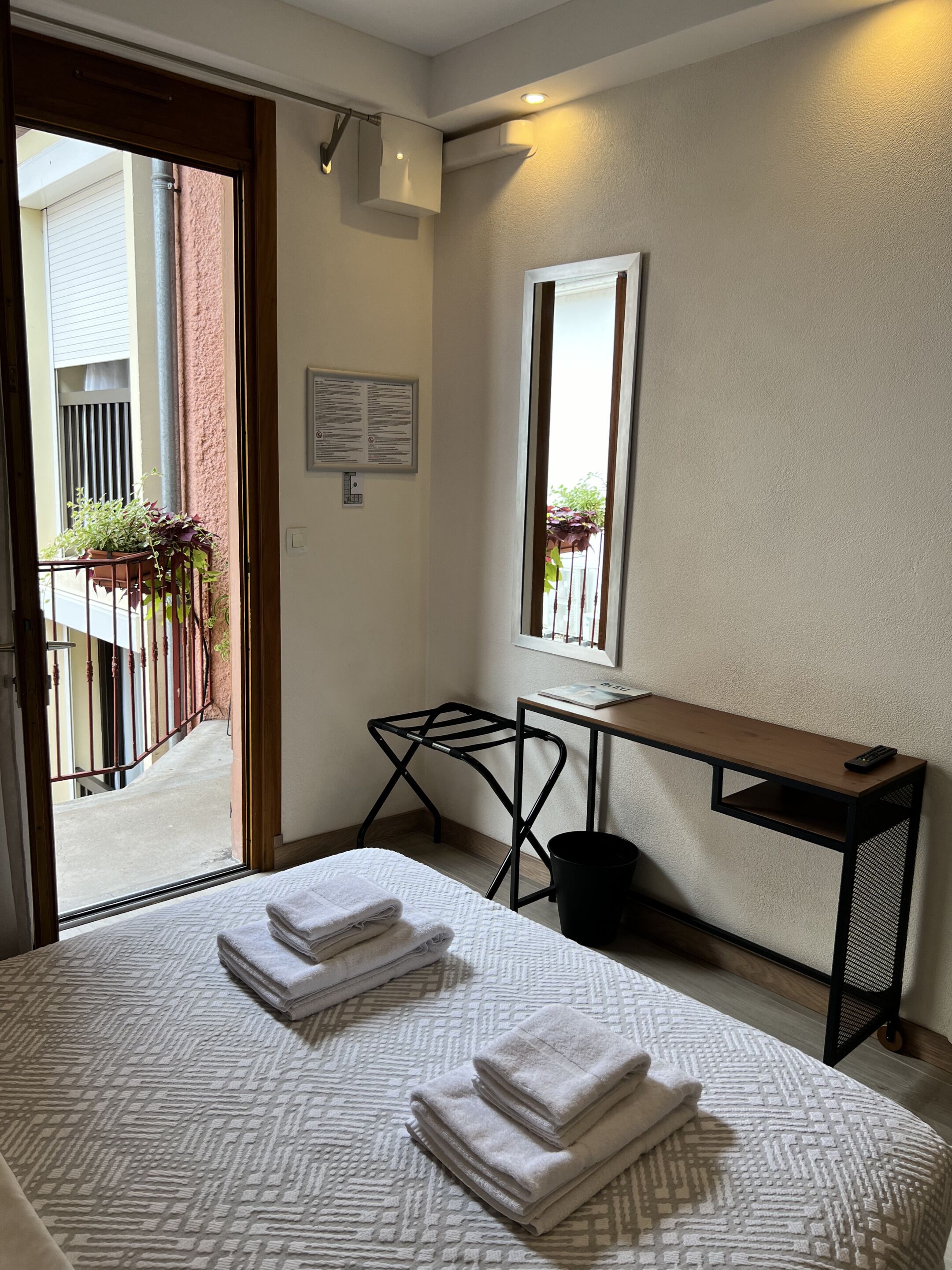 Habitación de hotel con cama, escritorio, silla, un cubo de basura pequeño y ventana con vistas al exterior.