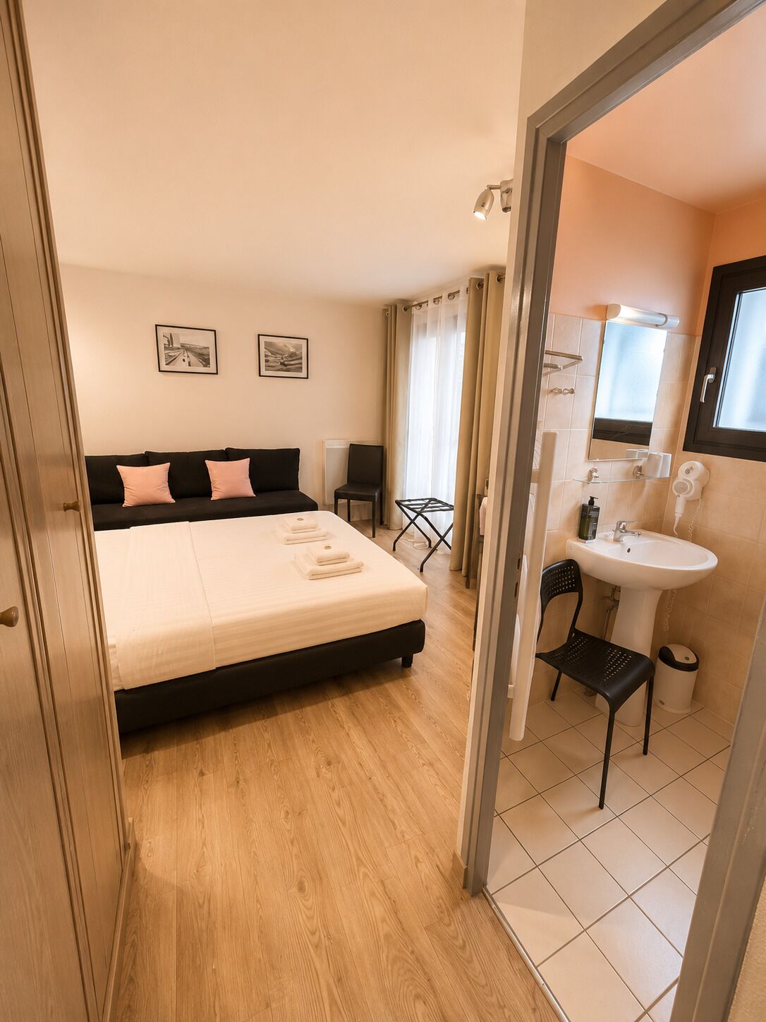 A bedroom with a bed, a bedside table, a chair, a window, and a bathroom with a sink and mirror.