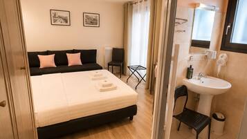 A bedroom with a bed, a bedside table, a chair, a window, and a bathroom with a sink and mirror.
