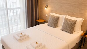 A hotel room with a large bed, a bedside table, a wall-mounted light, and a view through sheer curtains.