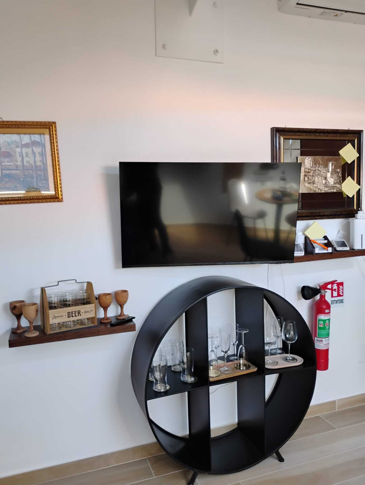 Une pièce comprenant un téléviseur fixé au mur, une étagère noire ronde avec des verres, une étagère en bois avec des verres et une caisse de bière, ainsi qu’un extincteur.