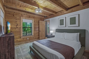 Ein Schlafzimmer mit Holzboden, einem Bett mit weißer Bettwäsche, einem Nachttisch mit Lampe und einem Fenster mit Blick auf Grünflächen.