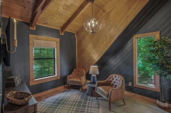 Ein gemütliches Zimmer mit hoher Decke, Holzbalken, einem Chandelier, zwei Ledersesseln, einem runden Tisch mit Lampe, einem Korbwäscher und einem großen Fenster mit Blick auf Bäume.