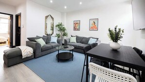 Un salon moderne avec un canapé gris, une table basse et une table à manger avec des chaises. Deux plantes en pot et deux tableaux encadrés ornent le mur.