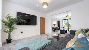 Ein modernes Wohnzimmer mit großem Flachbildfernseher an der Wand, einem grauen Sofa mit bunten Kissen, einem Glastisch und einer Topfpflanze.