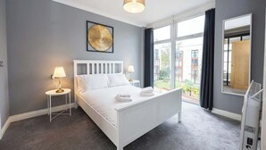Una habitación de hotel moderna con una cama blanca, un adorno redondo en la pared, una lámpara de noche y un amplio ventanal con vistas a edificios.