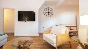 Una sala de estar moderna con un televisor de pantalla plana, un reloj de pared, una mesa de centro de vidrio, un sofá de mimbre y una mesa de comedor de madera.