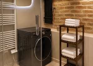 Une salle de bain moderne avec un lave-linge noir, une cabine de douche en verre, une étagère en bois où sont pliées des serviettes et un mur en briques.