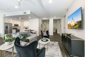 Un salon moderne comprenant un canapé, des fauteuils, une table basse et un coin repas avec une table et des chaises. On aperçoit, au fond, une cuisine équipée d’appareils électroménagers et de meubles de rangement.