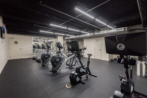 Une salle de sport équipée de divers appareils d’exercice, dotée d’un mur recouvert de miroirs et d’un plafond laissant apparaître les conduits.