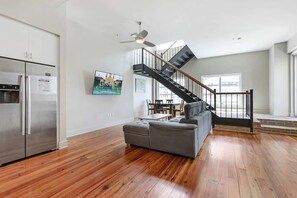 Ein modernes Wohnzimmer mit einem grauen Ecksofa, Holzboden, einem Edelstahlkühlschrank, einem flachbildfernseher an der Wand und einer Treppe mit schwarzer Geländer.