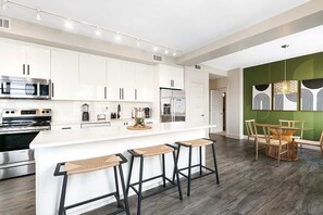 Una cocina moderna con gabinetes blancos, electrodomésticos de acero inoxidable, una isla central con taburetes y una zona de comedor con una pared de acento verde.