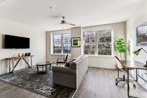 Un salon moderne avec un canapé gris, une petite table ronde, un bureau avec une lampe et un ventilateur de plafond.