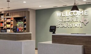 Ein Hotel-Empfangsbereich mit einem Schild, das 'Welcome to the Lone Star State' ankündigt, einem Selbstbedienungs-Check-in-Terminal und einer Minibar, gefüllt mit Snacks.