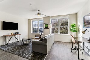 Un salon moderne avec un canapé gris, une petite table ronde, un bureau avec une lampe et un ventilateur de plafond.