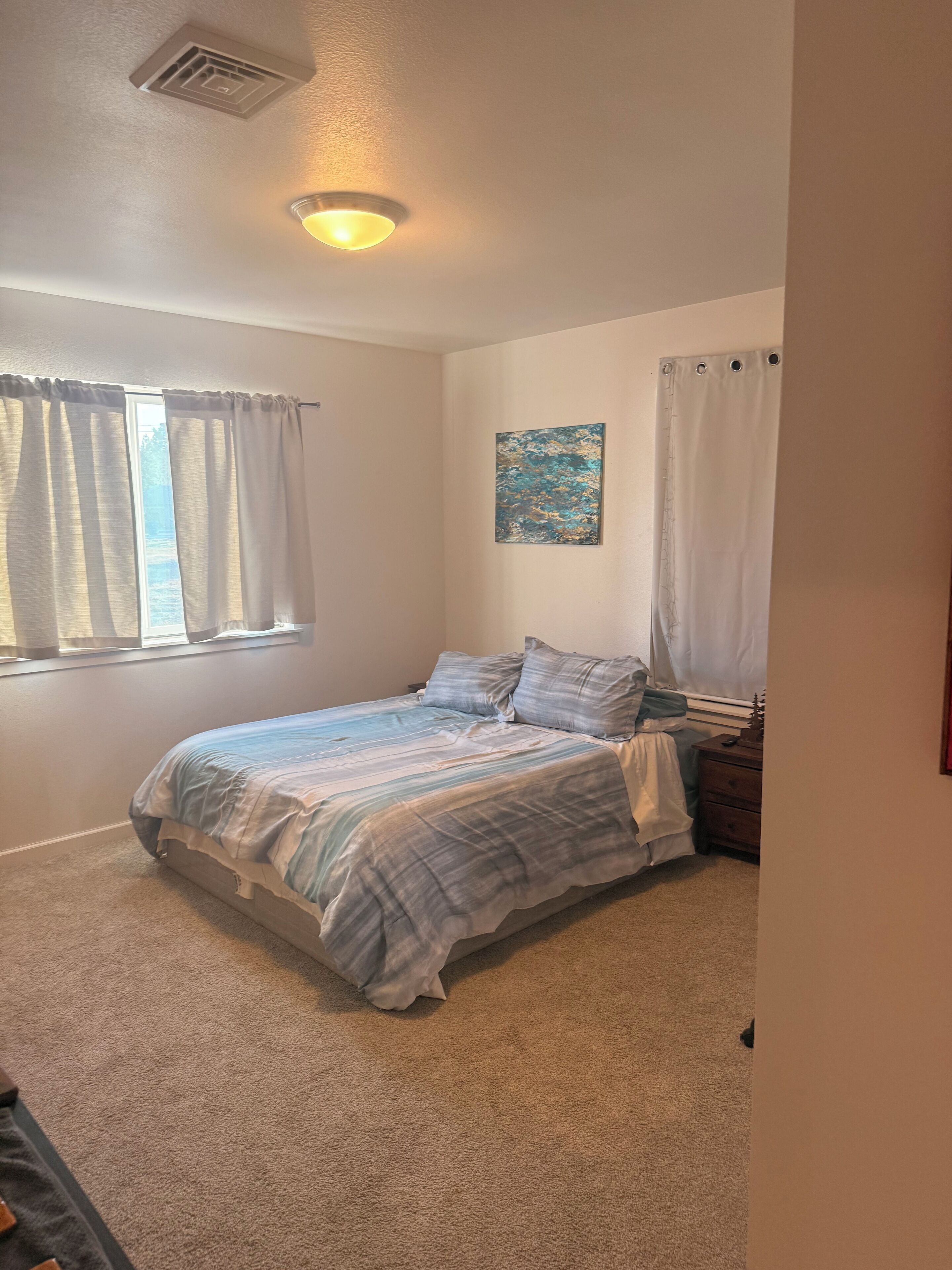 Une chambre à coucher avec un lit, une fenêtre avec des rideaux, une bouche d’aération au plafond et un tableau accroché au mur.