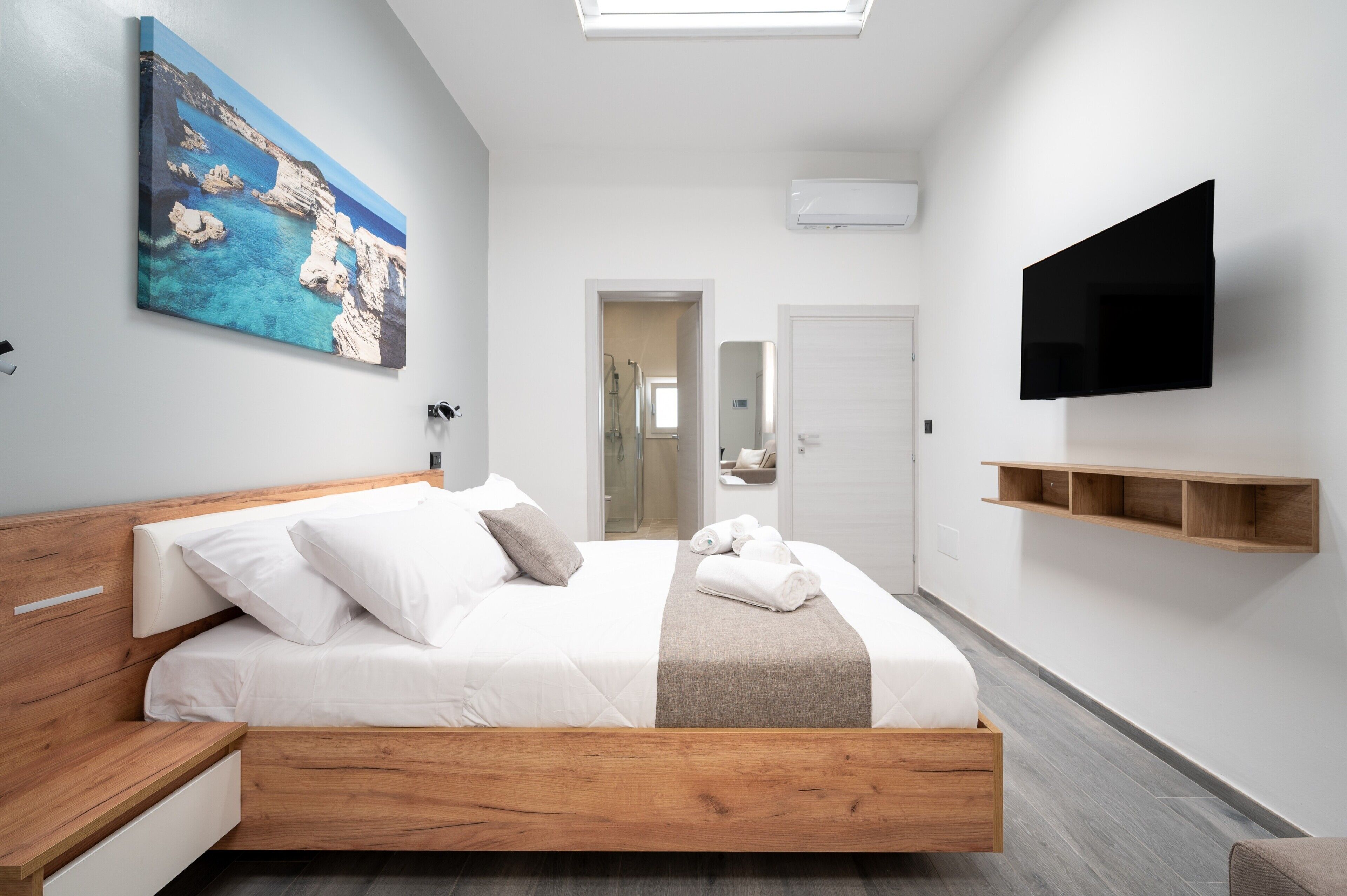 A modern hotel room with a wooden bed, a flat-screen TV, a painting, and a hallway leading to a bathroom.