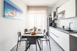 A modern kitchen with a dining area, a painting on the wall, and a window with sheer curtains. - Piazza Pertini - Room Smeraldo with Balcony, Wi-Fi and Air Conditioning (Melendugno)