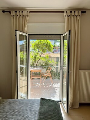 Ein Zimmer mit Bett, einem Fenster mit Vorhängen, einem Blick auf einen Balkon mit Tisch und Stühlen sowie einer Heizung.