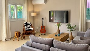 Una sala de estar con un sofá gris, una mesa de centro de madera, una guitarra, una lámpara y un televisor montado en la pared.