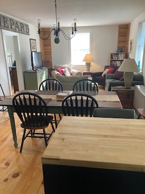 A dining area with a wooden table and chairs, a television on a cabinet, a chandelier, and a bookshelf in the background.