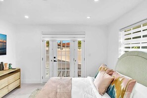 Ein Schlafzimmer mit Bett, Kommode, Fenster mit Ausblick und einer Tür, die auf einen Balkon führt.