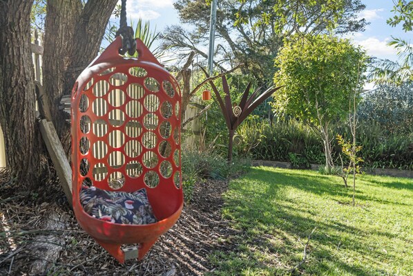 Una hamaca colgada entre árboles en un jardín con una valla de madera y abundante vegetación al fondo.