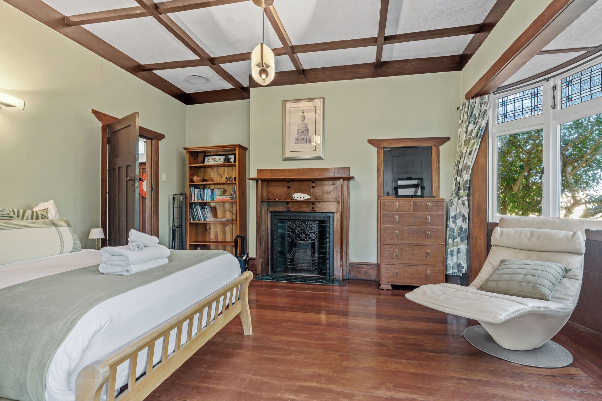 A room with a bed, a wooden bookshelf, a fireplace, a wooden cabinet, and a window with a view of trees.