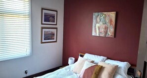 A bedroom with a bed, a window with blinds, and two framed pictures on the wall.