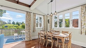 A dining area with a wooden table and chairs, large windows, and a view of greenery outside.