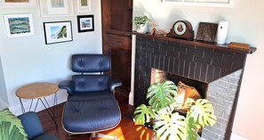 A room with a black leather chair, a small round table, a fireplace with a brick surround, and framed pictures on the wall.
