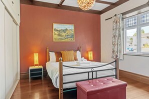 A bedroom with a bed, bedside lamps, a red ottoman, a painting on the wall, and a window with curtains.