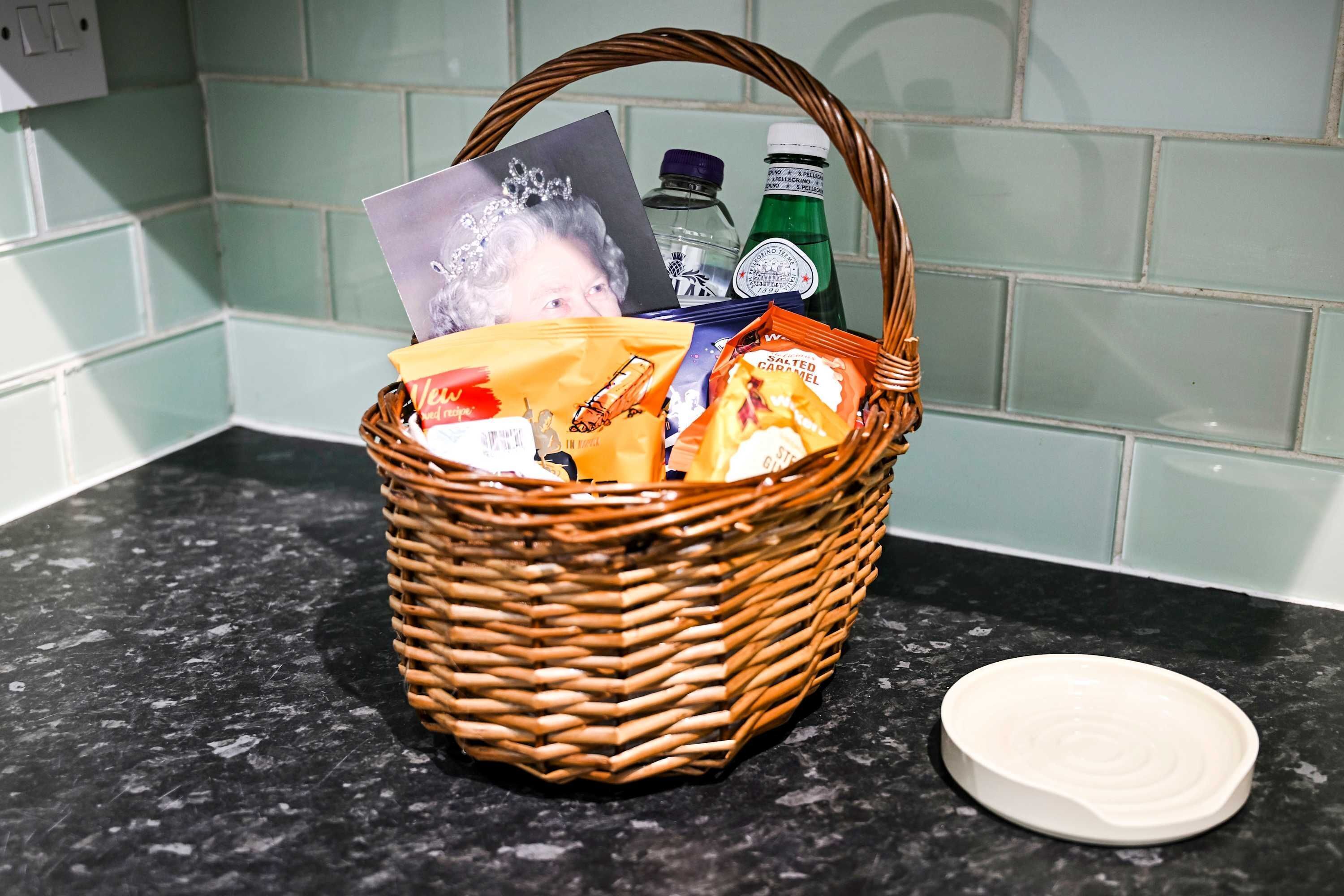 A wicker basket on a kitchen counter with a white lid, containing various items including a photo, a water bottle, and snacks.