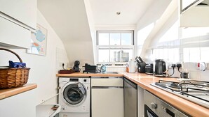 A compact kitchen with a washing machine, dishwasher, stove, and a large window providing natural light. - Chelsea Loft - Stylish Sanctuary (London)