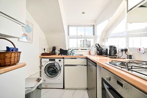 A compact kitchen with a washing machine, dishwasher, stove, and a large window providing natural light. - Chelsea Loft - Stylish Sanctuary (London)