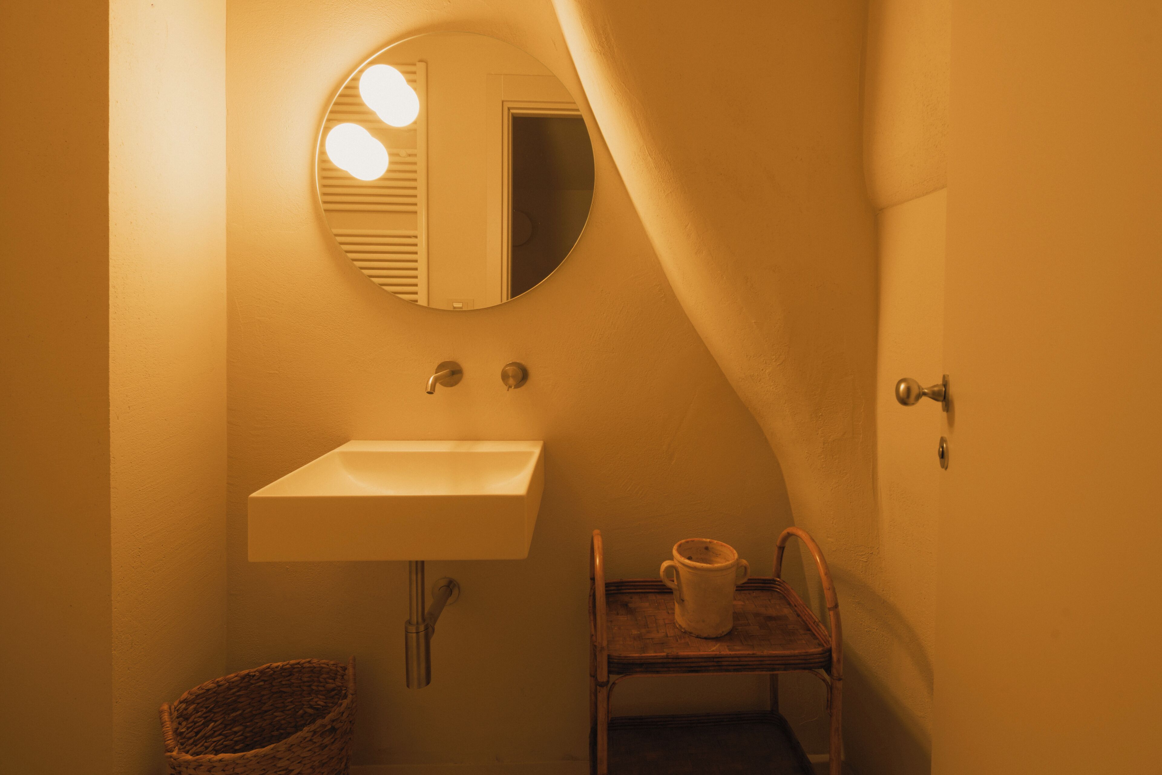 A bathroom with a round mirror, a sink, a small shelf with a cup, and a wicker basket.