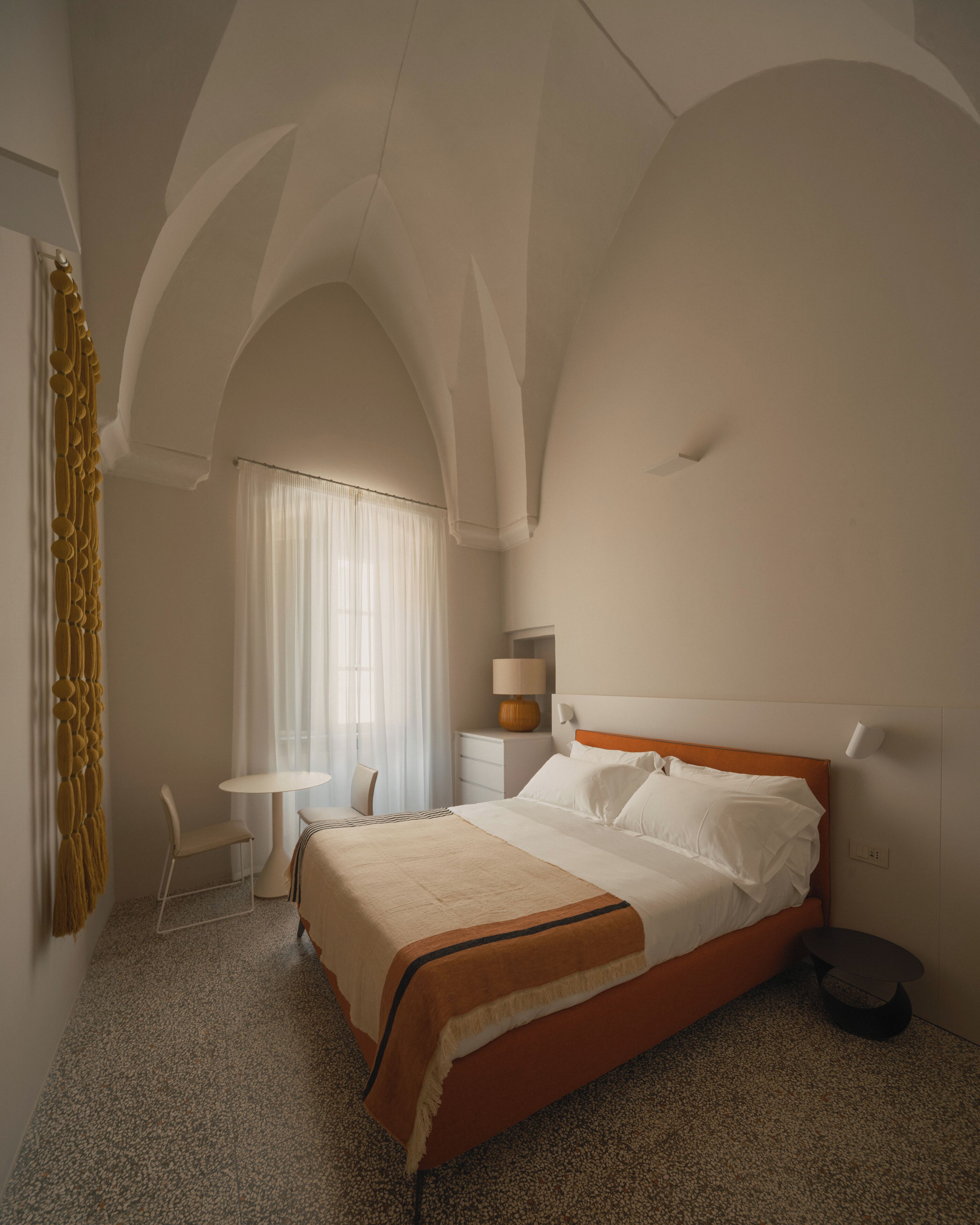 Un dormitorio con cama, mesita de noche, silla, lámpara y ventana con cortinas.