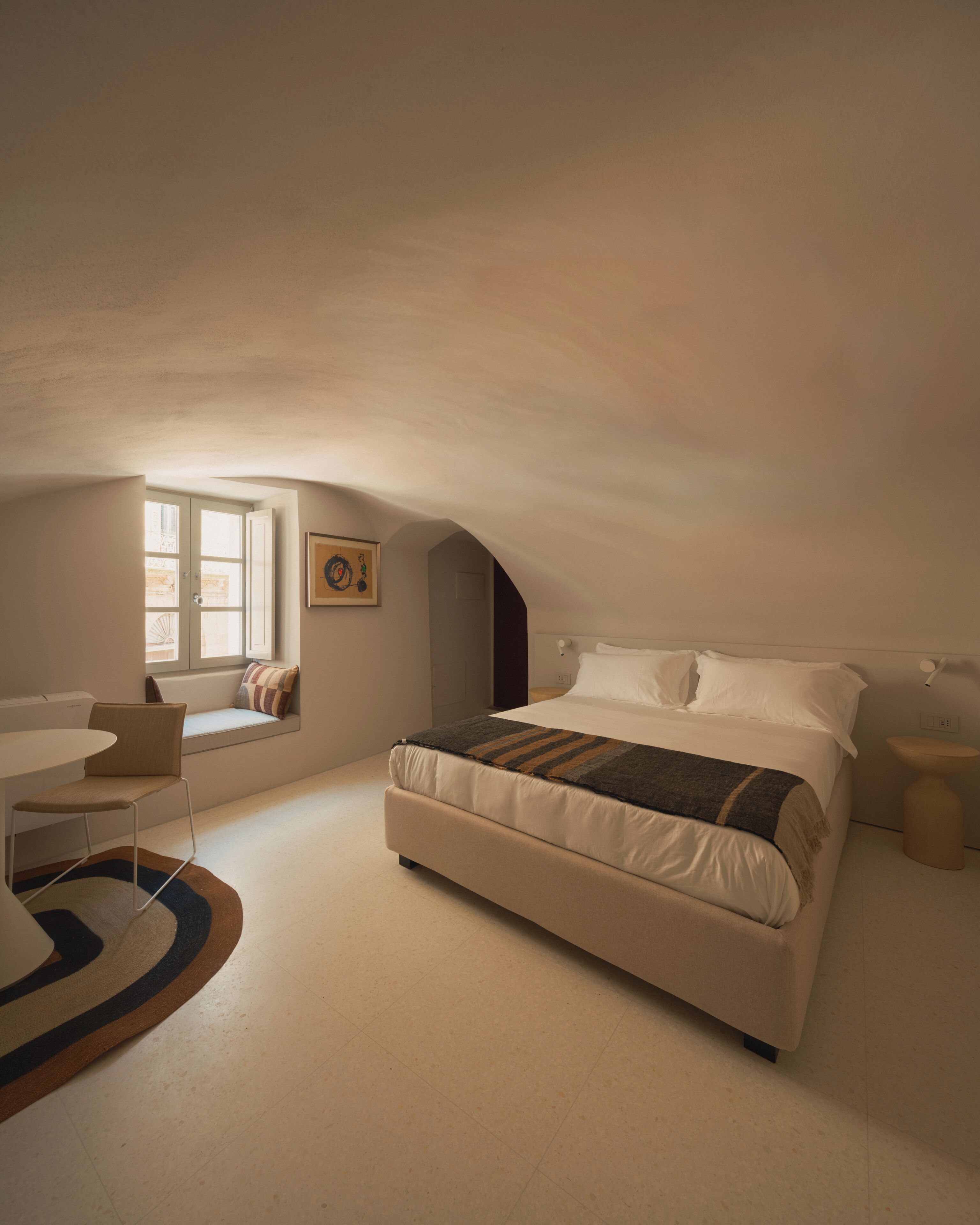 A bedroom with a bed, a window seat, a desk, and a small table.