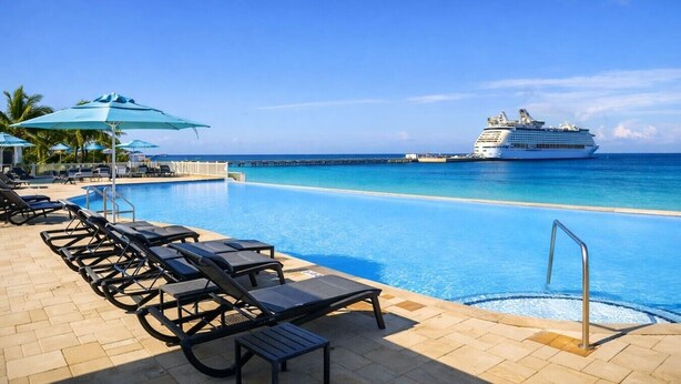 Un área junto a la piscina con sillones y sombrillas, un muelle que se adentra en el océano, y un crucero amarrado cerca.