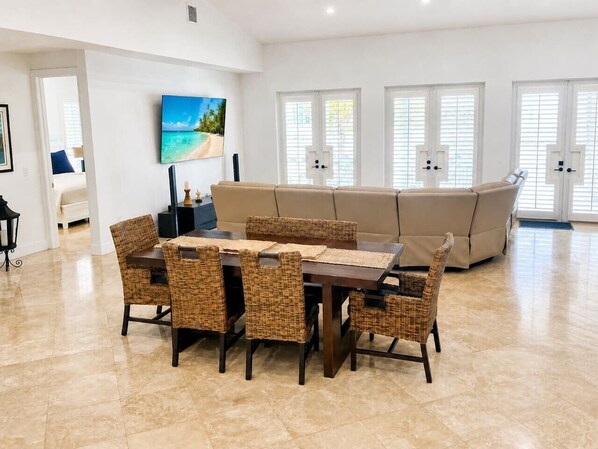 A spacious living area with a dining table, wicker chairs, a beige sofa, and a television mounted on the wall.