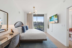 A hotel room with a bed, a desk with a lamp, a large window with curtains, a painting, and a television. - Stylish Wynwood Apartment with Resort Pool and Gym (Miami)