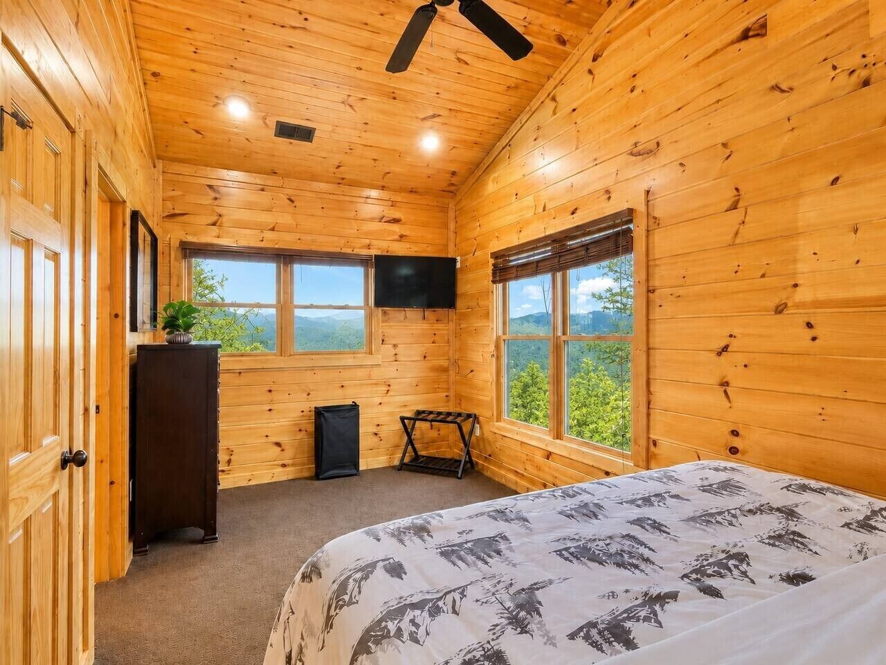 A wooden cabin room with a bed, a TV, a small table, and a window with a view.