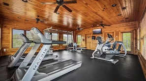 Ein Fitnessraum mit Laufbändern, Ergometer und Krafttrainingsgeräten in einer Holzhütte mit Holzwänden und -decke.