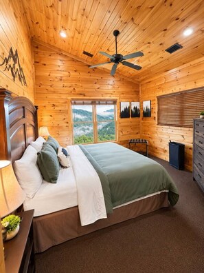 Ein Schlafzimmer einer Holzhütte mit einem Bett, einem Deckenventilator, einem Fenster mit Blick auf die Berge und einer Kommode.