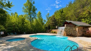 A swimming pool with a waterfall feature, surrounded by stone tiles and trees. - Views Forever | Smoky Mountain Views, Hot Tub, Theater & Game Room (Gatlinburg)