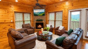 A cozy log cabin interior with leather sofas, a fireplace, and a television mounted on the wall. - Views Forever | Smoky Mountain Views, Hot Tub, Theater & Game Room (Gatlinburg)