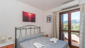 Un dormitorio con cama, mesita de noche, aire acondicionado instalado en la pared, ventana con cortinas e imagen en un marco.