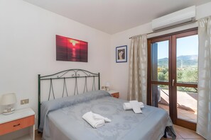 Ein Schlafzimmer mit Bett, Nachttisch, Wandklimaanlage, Fenster mit Vorhängen und einem gerahmten Bild.