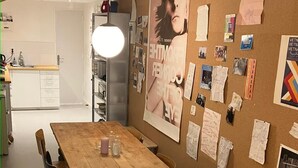 Uma área de refeições com mesa de madeira, cadeiras e um mural de cortiça com diversos papéis e fotografias.