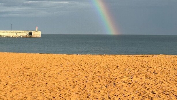 Una playa con un arcoíris sobre el agua y un faro a lo lejos.